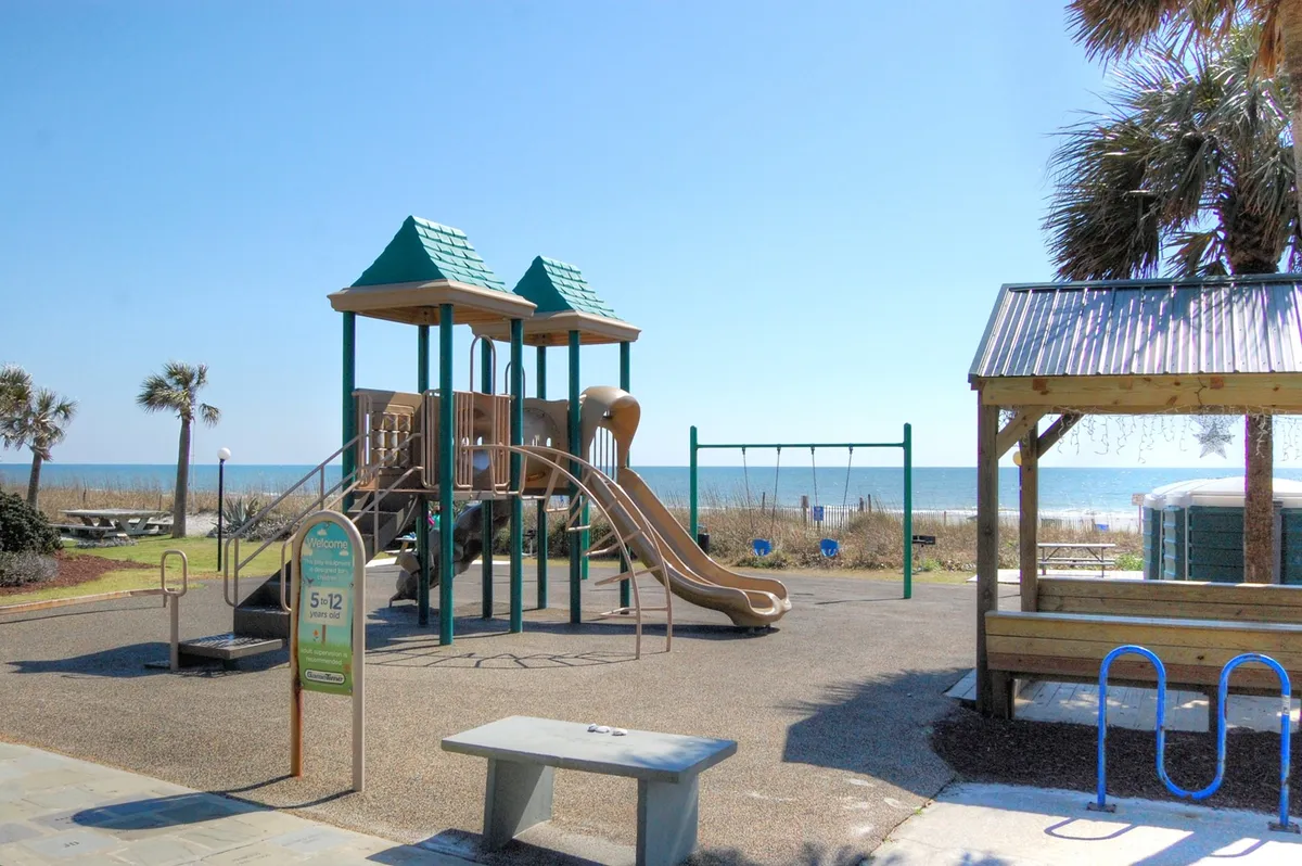This energetic exterior shot features a playground structure and swings set against a picturesque beach backdrop with palm trees. Children are playing, and families are enjoying picnics nearby, with the sound of the ocean in the air. The setting exudes joy and family bonding, highlighting the variety of activities available just steps from the property.