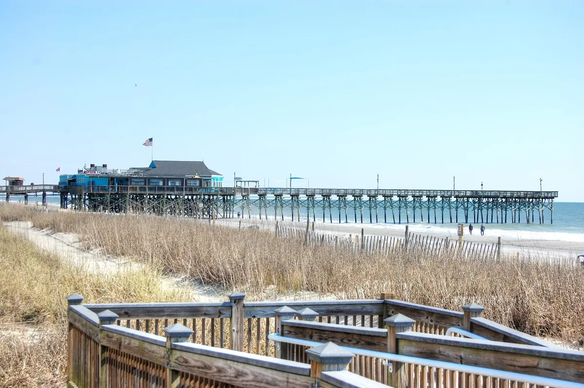 This image showcases a classic pier extending over the ocean, with golden sand dunes and beach grass in the foreground. The tranquil waters and expansive sky hint at the proximity of the beach and the allure of sunrise or sunset strolls.