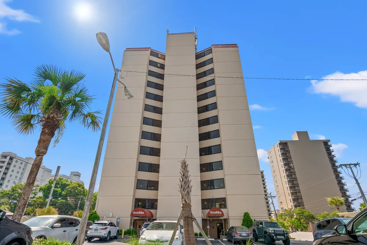 This image shows the exterior of a tall condo building lined with palm trees and a visible parking area. The setting is bright and welcoming, evoking the feel of a modern, well-maintained coastal residence. The outdoor area and the facade suggest a lively yet peaceful environment suitable for long-term stays.