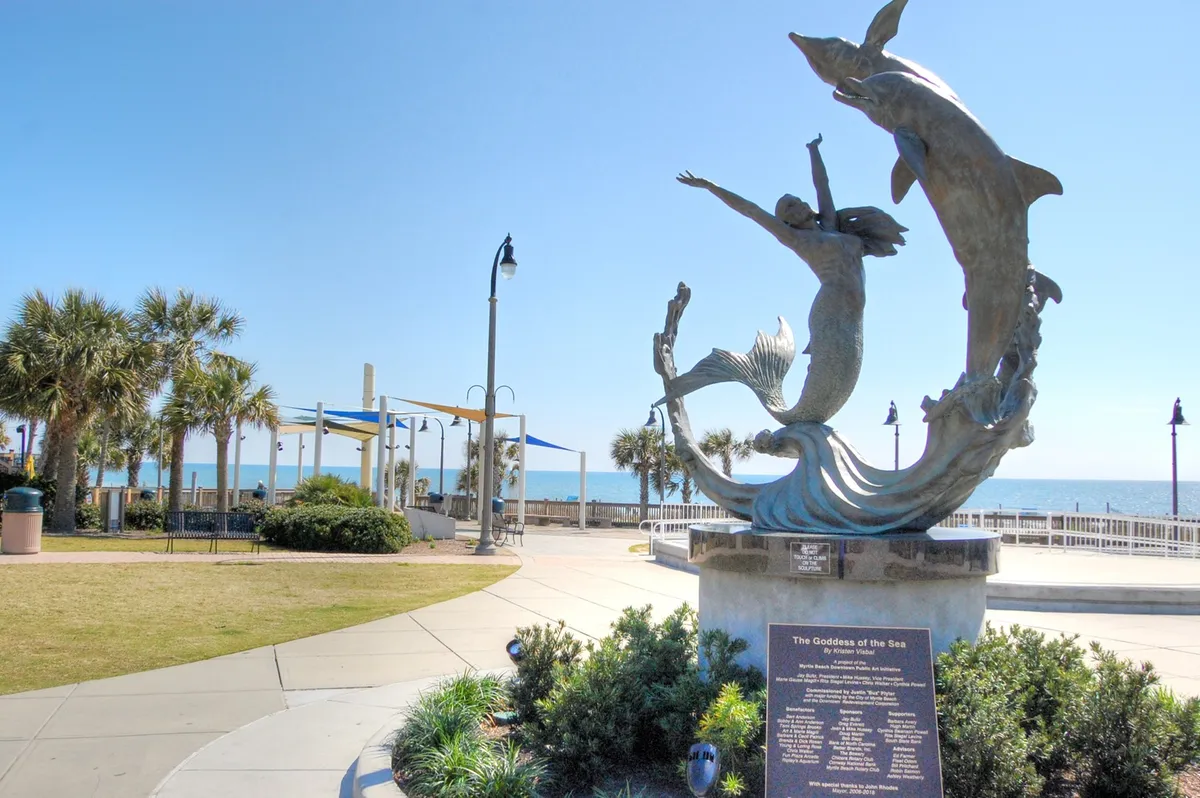 An uplifting seaside scene featuring the iconic Goddess of the Sea sculpture, ocean views, and palm trees. The promenade is perfect for leisurely walks, with the sun setting over the ocean and a sense of local culture and relaxation.