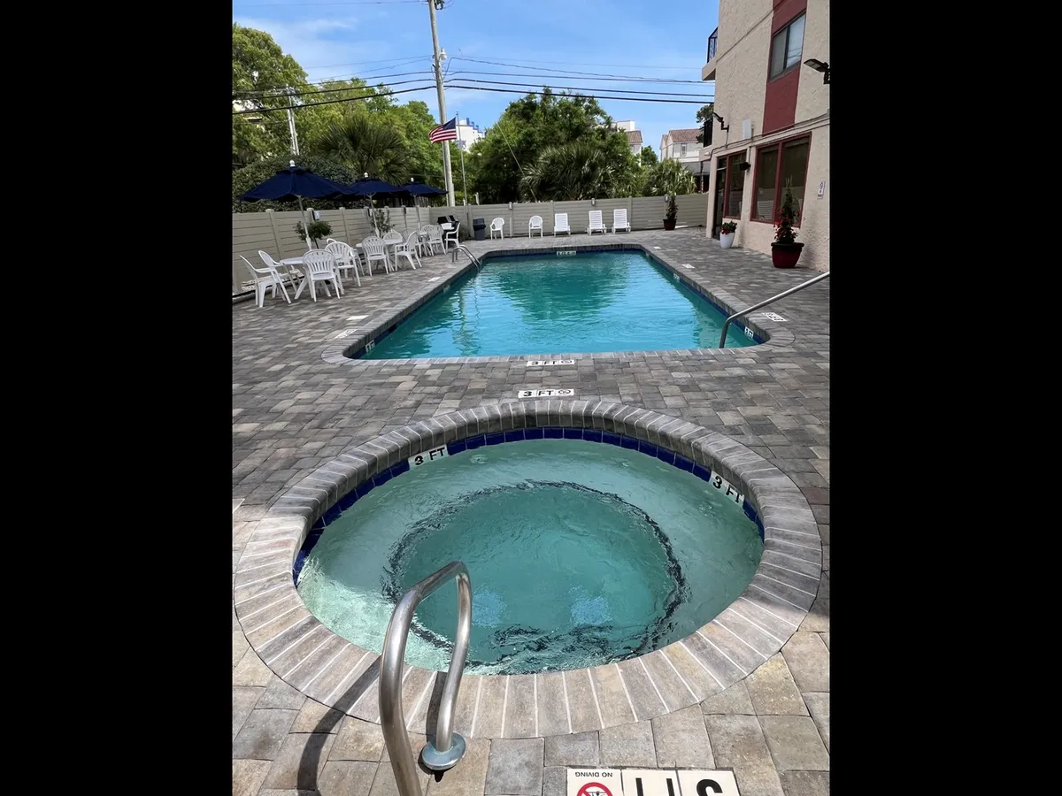 A panoramic view of a sparkling pool area with a hot tub, shaded lounge chairs, and large umbrellas. The pool deck is designed for leisure, offering spaces for swimming, unwinding in the hot tub, and enjoying the Myrtle Beach sunshine in a social setting.
