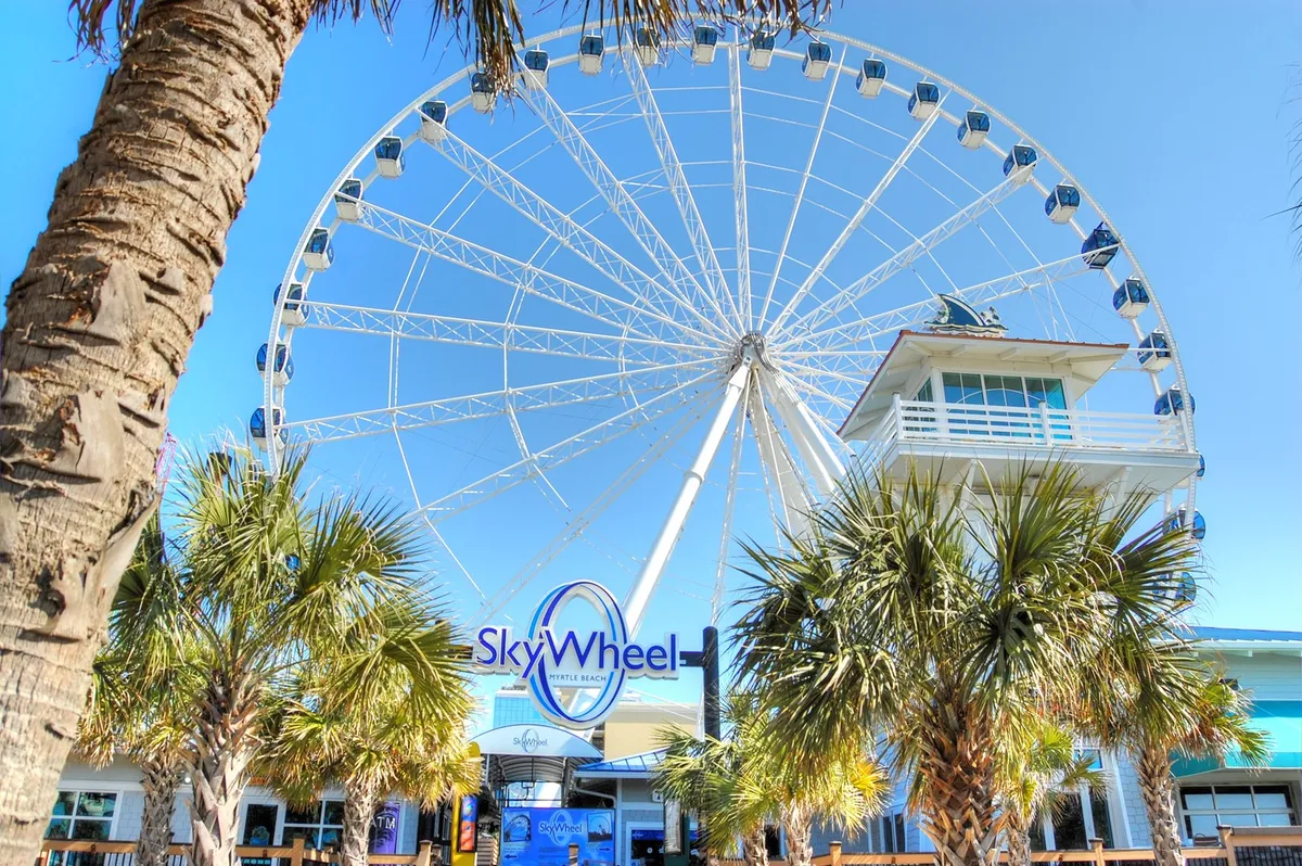 This image captures the iconic Myrtle Beach SkyWheel, framed by palm trees and the entrance building bustling with visitors. The vibrant atmosphere and clear coastal sky evoke the excitement of enjoying one of Myrtle Beach’s most popular attractions.