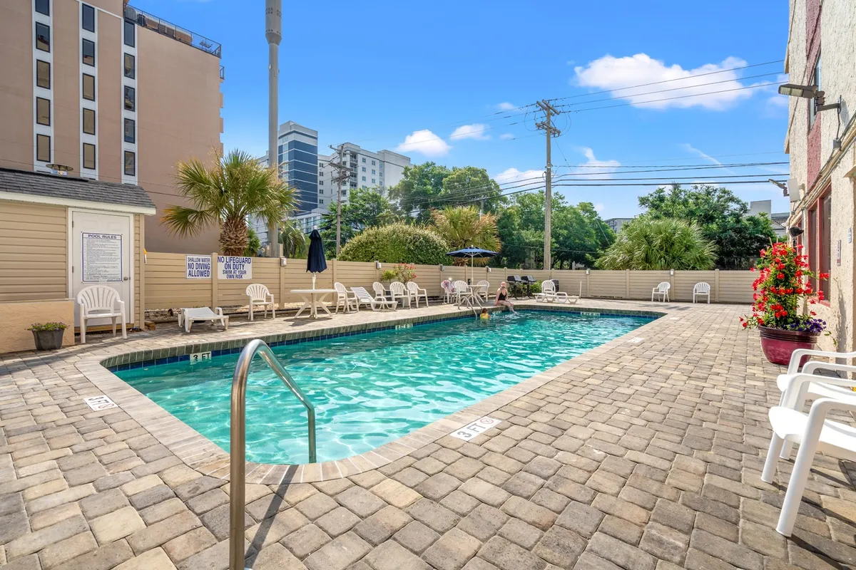 A sun-drenched outdoor swimming pool bordered by lounge chairs and lush palm trees, with inviting clear water that suggests a relaxing, resort-like atmosphere. The surrounding greenery creates a tranquil backdrop, highlighting the space as a central amenity for residents and guests.