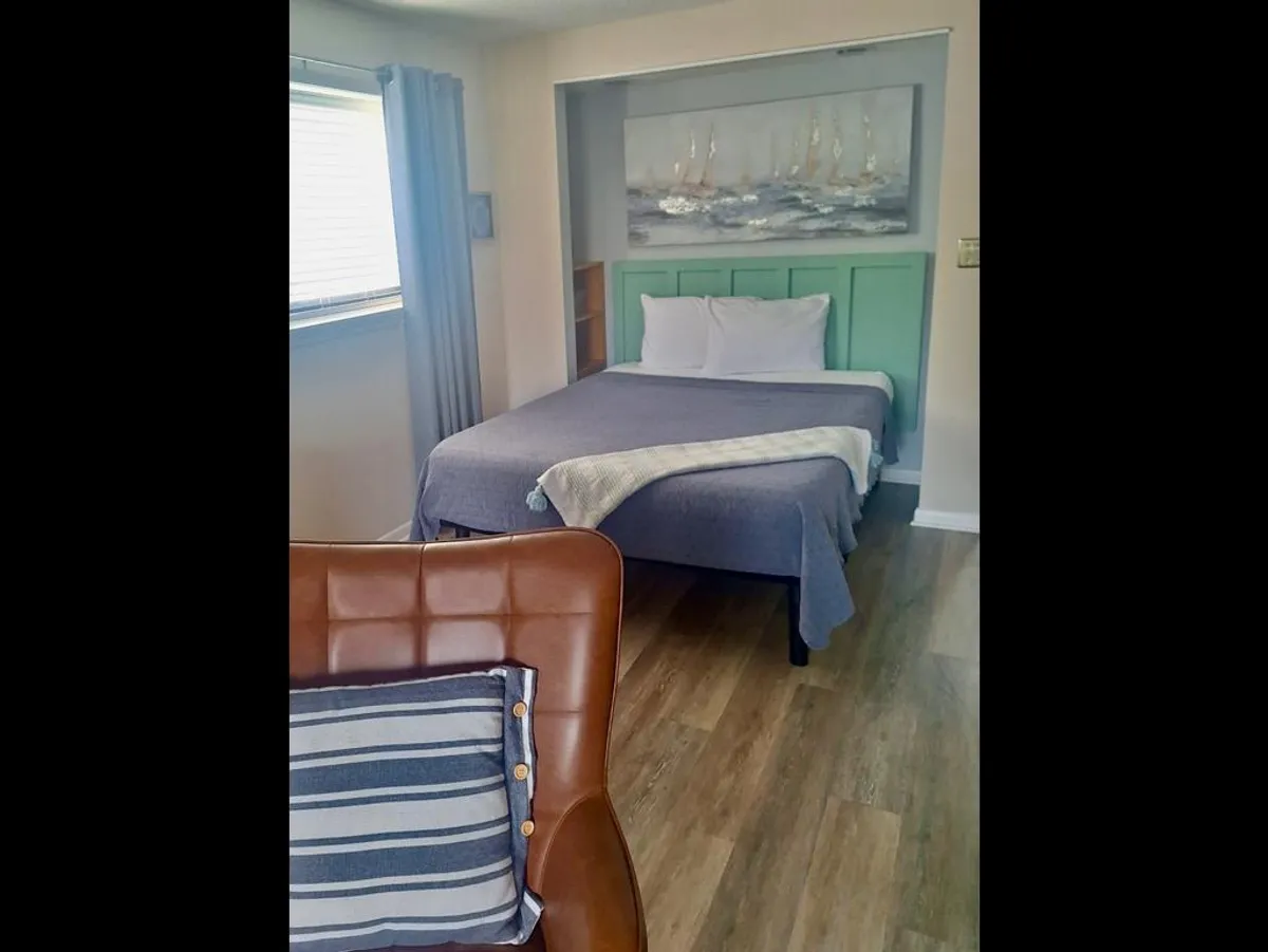 A tranquil bedroom scene featuring a plush bed with crisp linens, a painting of sailboats above the headboard, and a comfortable chair with a striped pillow positioned nearby. The decor is calm and coastal, with soft lighting and an inviting atmosphere.