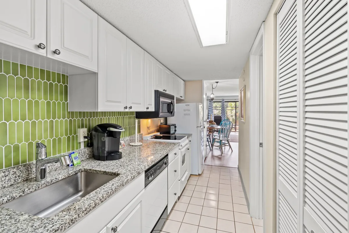 A bright kitchen featuring a granite countertop, sleek green backsplash, modern appliances, and a view into the adjacent dining area. The space exudes a cheerful atmosphere, with plenty of work surface and amenities for cooking and entertaining.