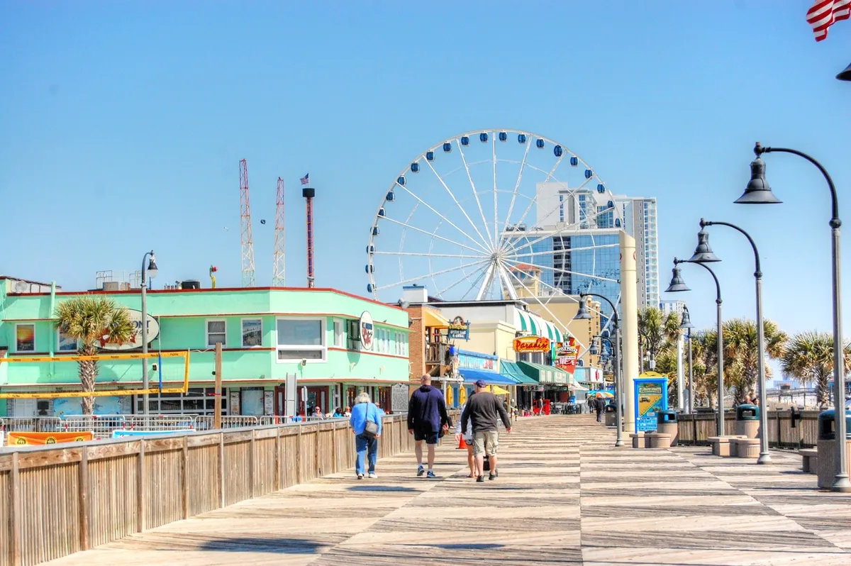 A lively boardwalk scene featuring the Ferris wheel, colorful buildings, and crowds of people enjoying amusement rides and shopping. The joyful, relaxed mood captures the spirit of exploration and fun along Myrtle Beach’s famous beachfront.