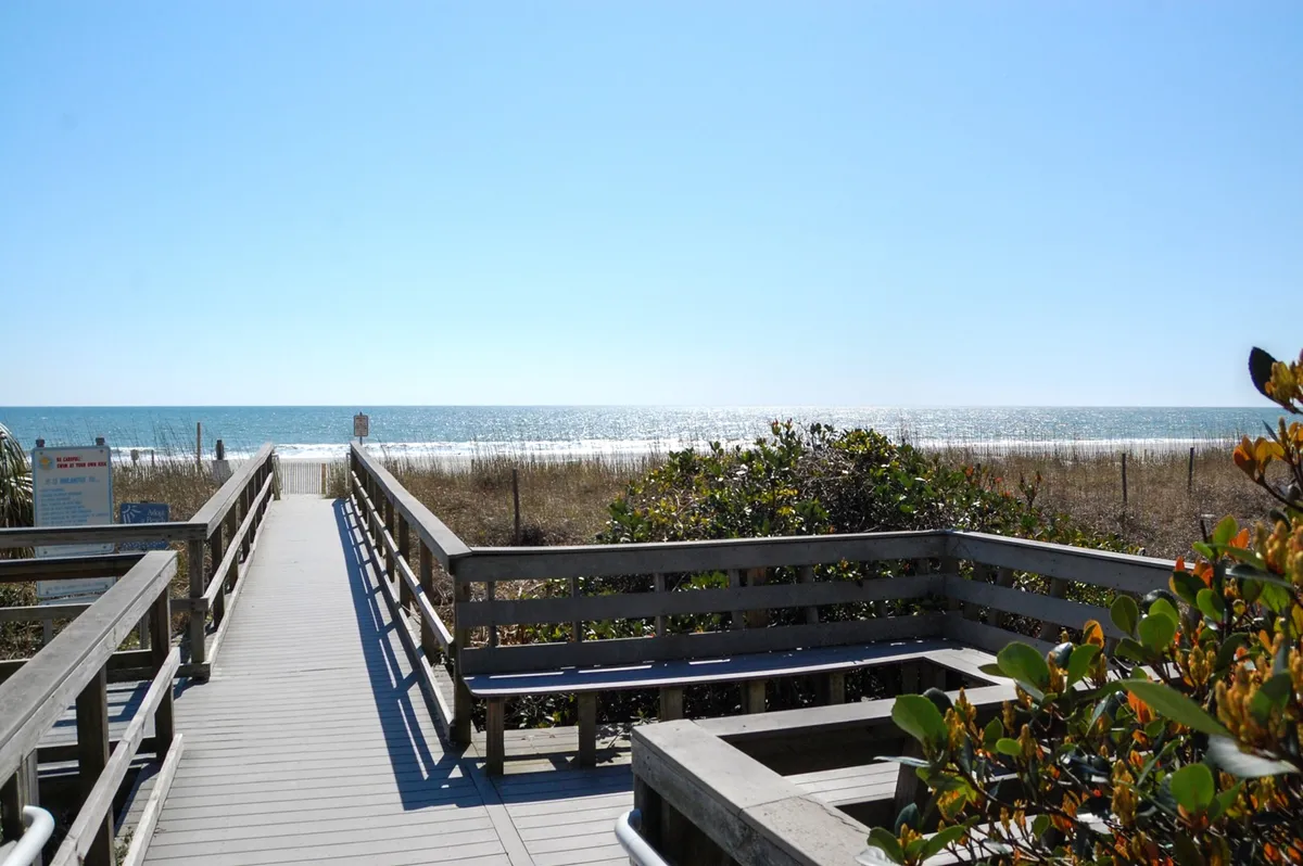 A picturesque wooden boardwalk leads through dune grasses toward the ocean, with tranquil blue water stretching beyond. The inviting pathway hints at effortless beach access and the chance to enjoy both sunrise and sunset vistas.