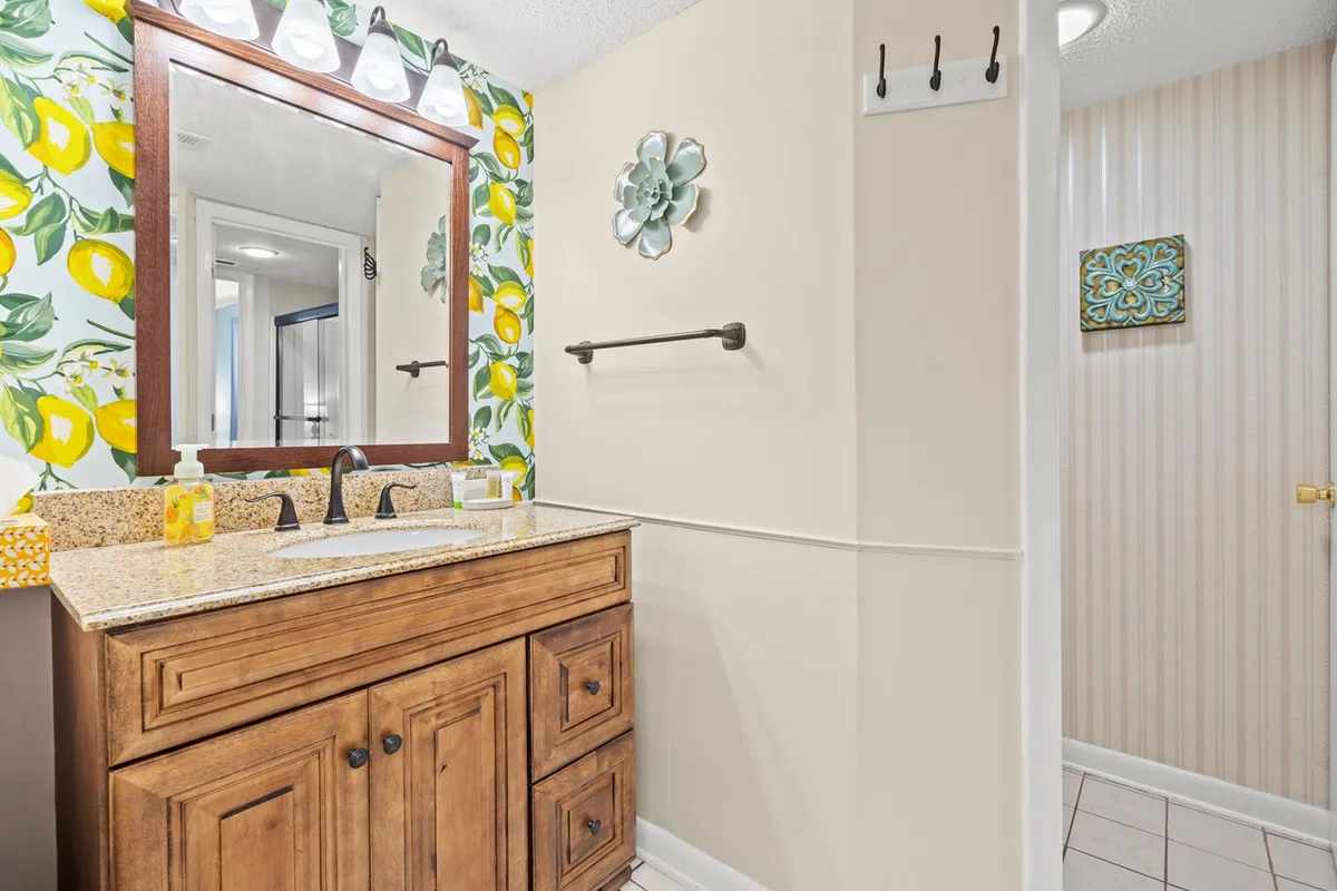A cheerful half-bathroom featuring a vanity with a granite countertop, lemon-themed wallpaper, a framed mirror, and playful decorative wall art. The space is bright and full of character, perfect for quick refreshes and added convenience.